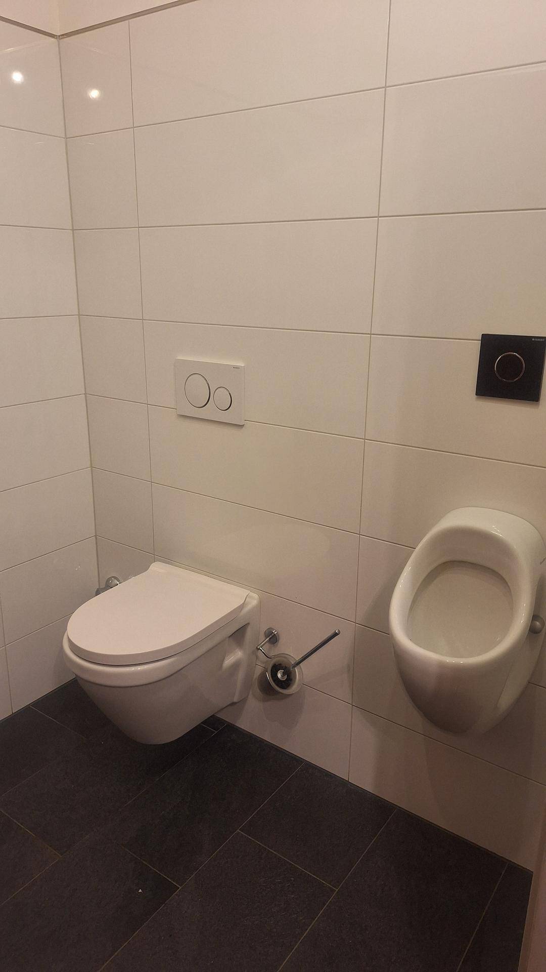 Separates WC mit wandhängender Toilette und Urinal, ausgestattet mit dunklen Bodenfliesen.