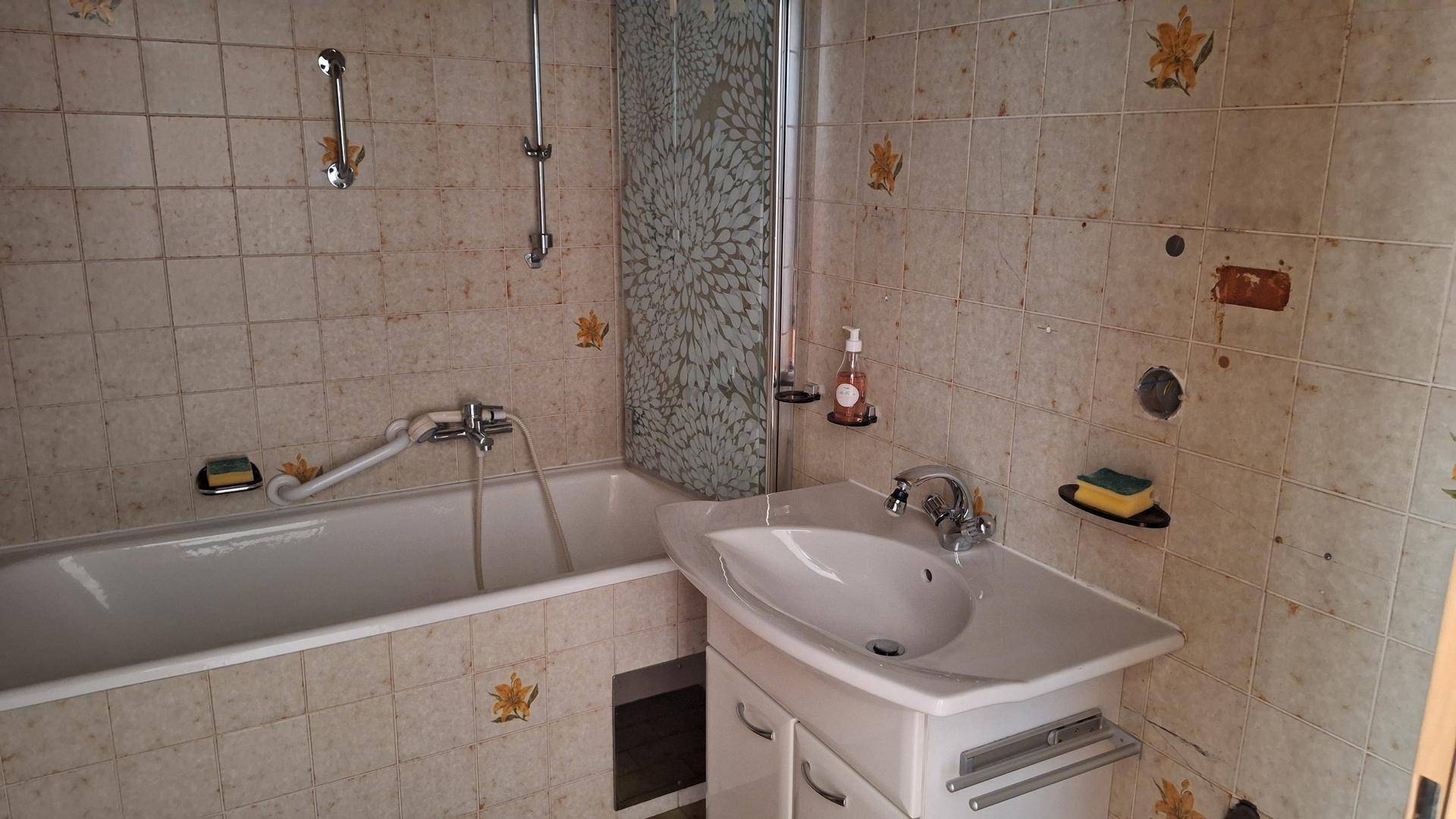 Badezimmer im 80er-Jahre-Stil mit Badewanne, Waschbecken und beigen Fliesen mit Blumenmuster.