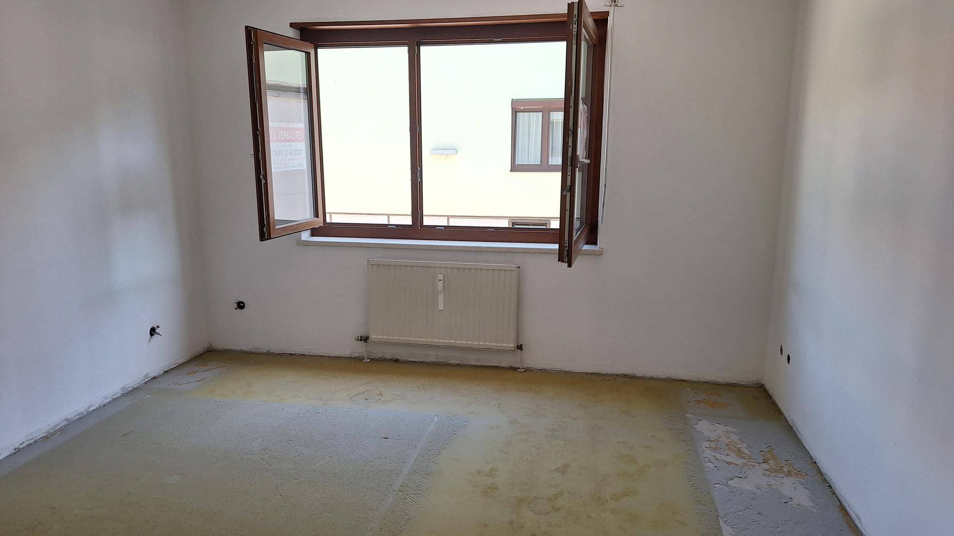 Heller Raum mit Fenster und Heizkörper, der einen Blick auf ein gegenüberliegendes Gebäude ermöglicht.