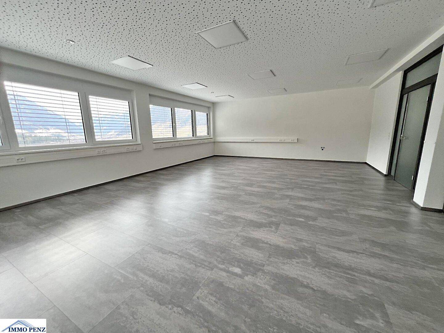 Leerer, heller Büroraum mit großen Fenstern, modernen Bodenfliesen und Blick auf die umliegende Landschaft.