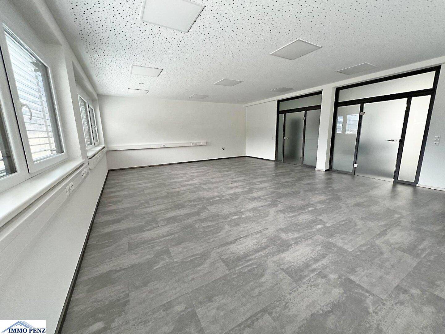 Geräumiger, leerer Büroraum mit großen Fenstern, modernen Bodenfliesen und Glastüren zu weiteren Bereichen.