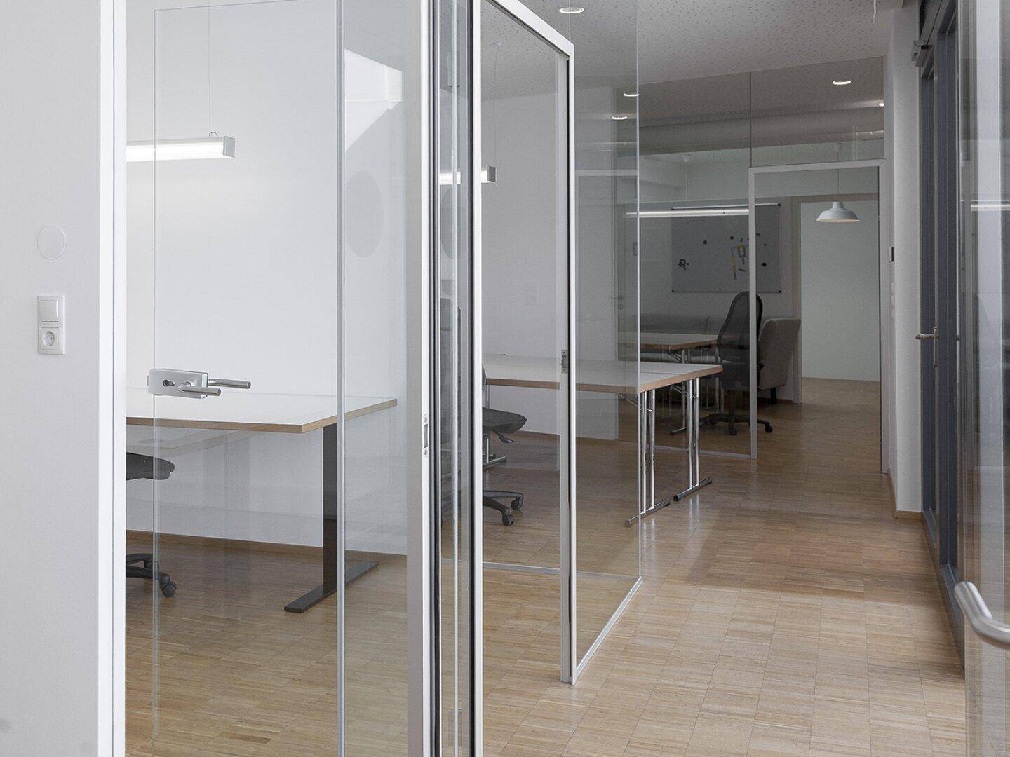 Moderner Bürobereich mit Glaswänden, Holzboden und viel Tageslicht.