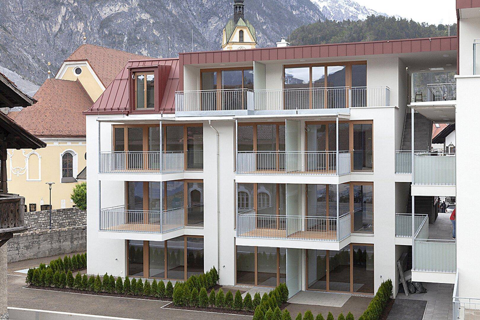 Moderne Wohnanlage mit mehreren Balkonen und Blick auf die umliegende Berglandschaft und Kirche.