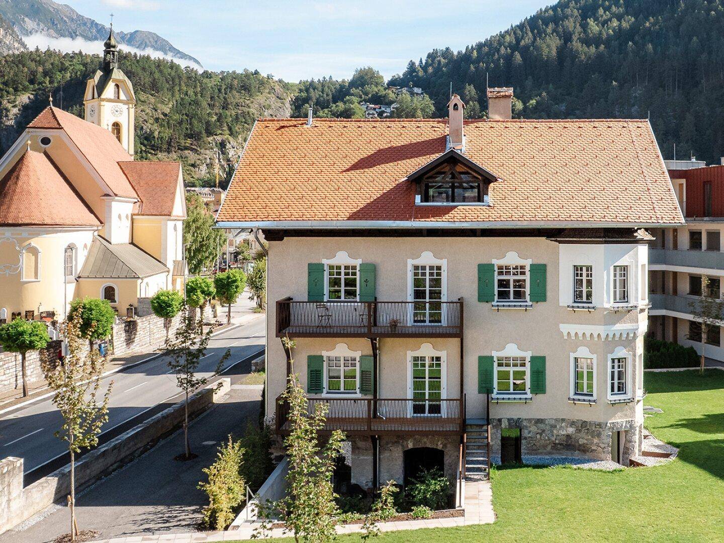 Historisches Gebäude mit grünen Fensterläden, Balkonen und Bergblick in ländlicher Umgebung.