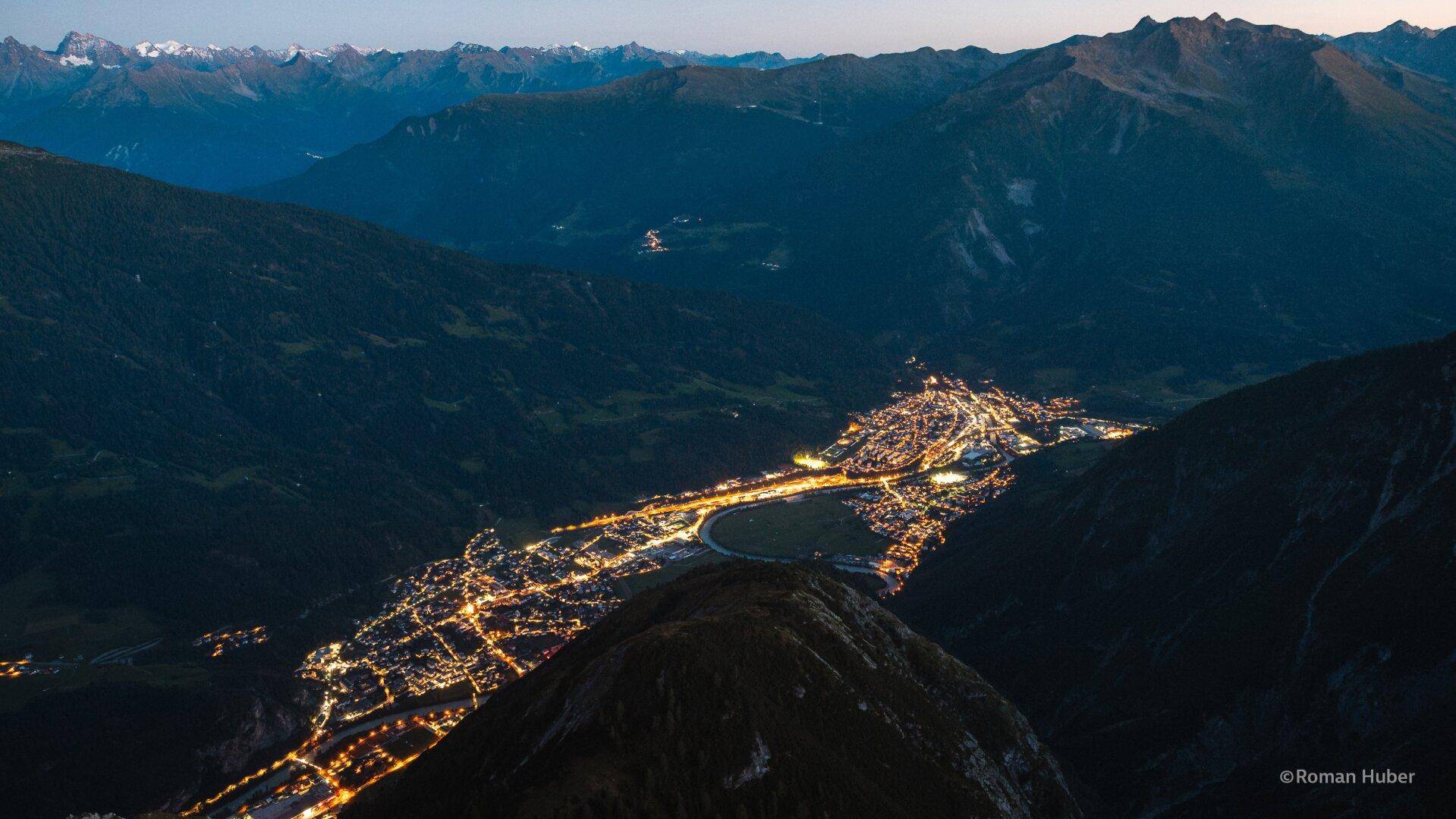 Nachtaufnahme einer beleuchteten Stadt im Tal, umgeben von dunklen Bergketten unter klarem Himmel.