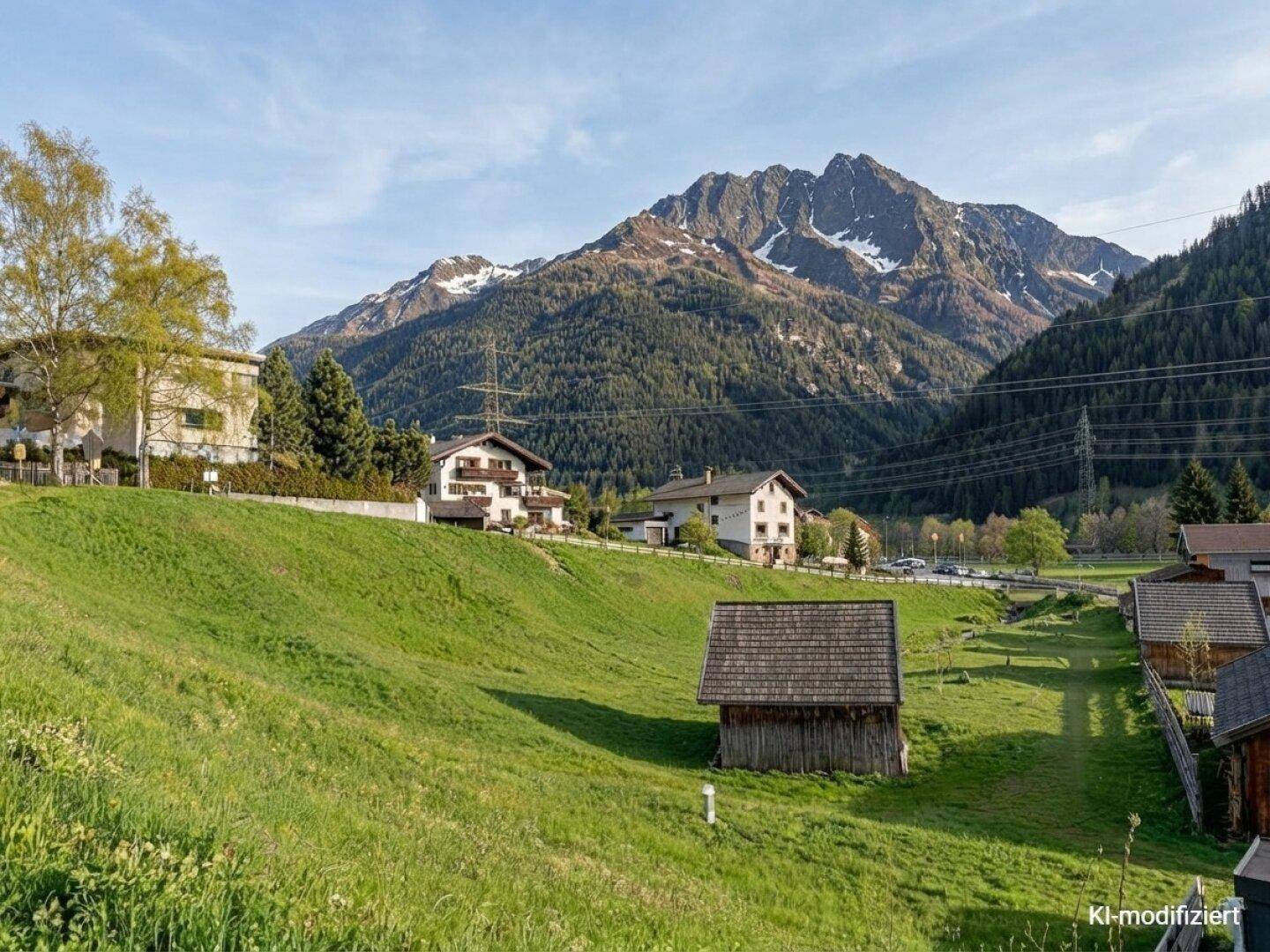 Grüne Wiese mit kleinem Holzschuppen und majestätischen Bergen im sonnigen Hintergrund.