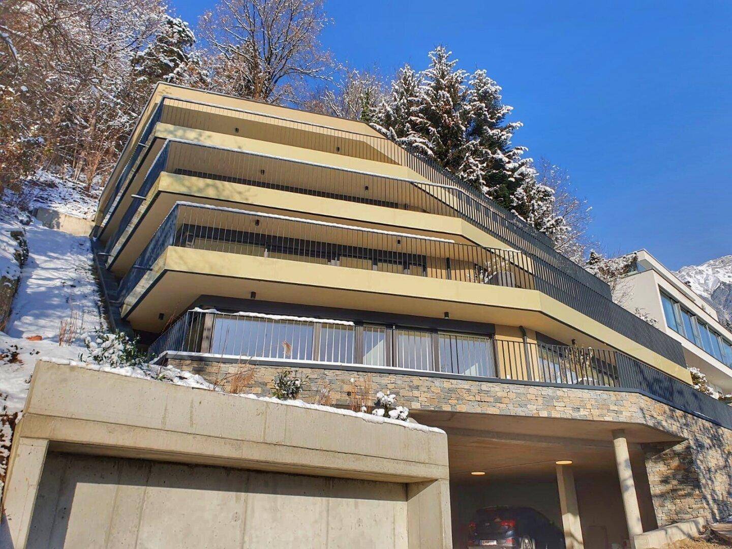 Modernes Apartmentgebäude in Hanglage mit mehreren Balkonen und Tiefgaragenzufahrt im Winter.