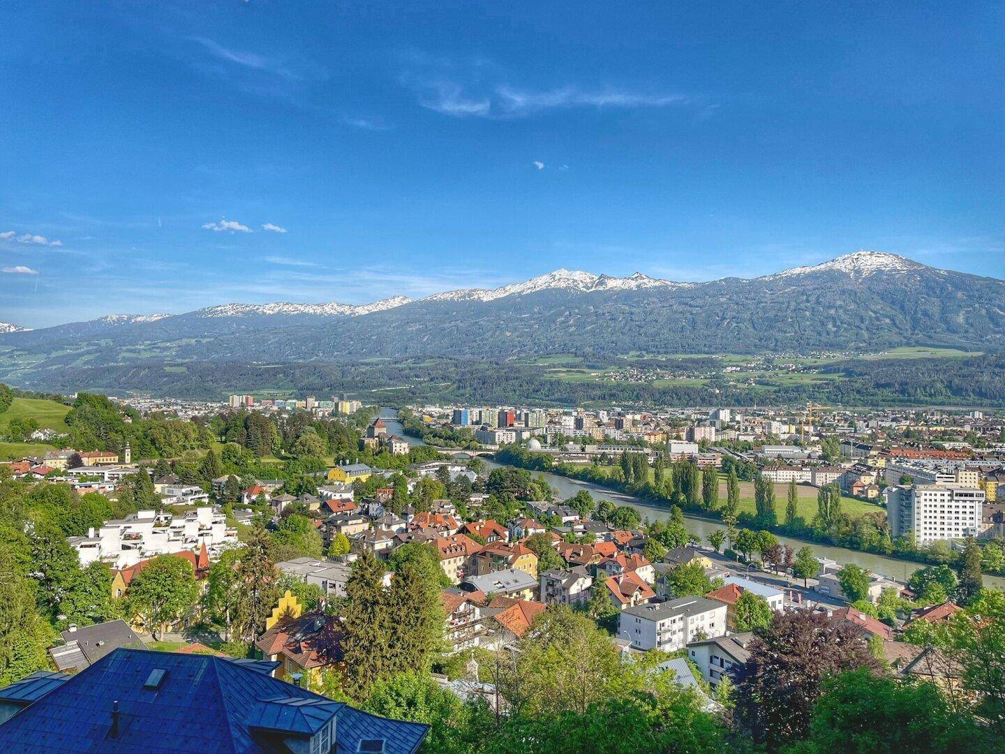Weitläufiger Panoramablick über eine Stadt mit Fluss, grünen Hügeln und schneebedeckten Bergen.