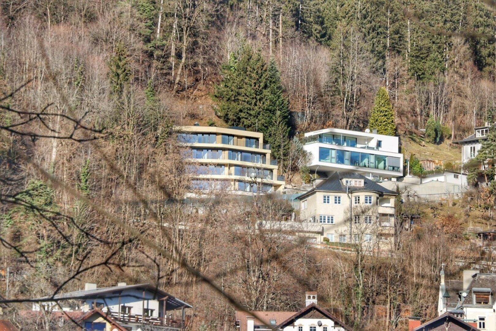 Panoramablick auf mehrere moderne und traditionelle Gebäude in einer bewaldeten Hanglage.