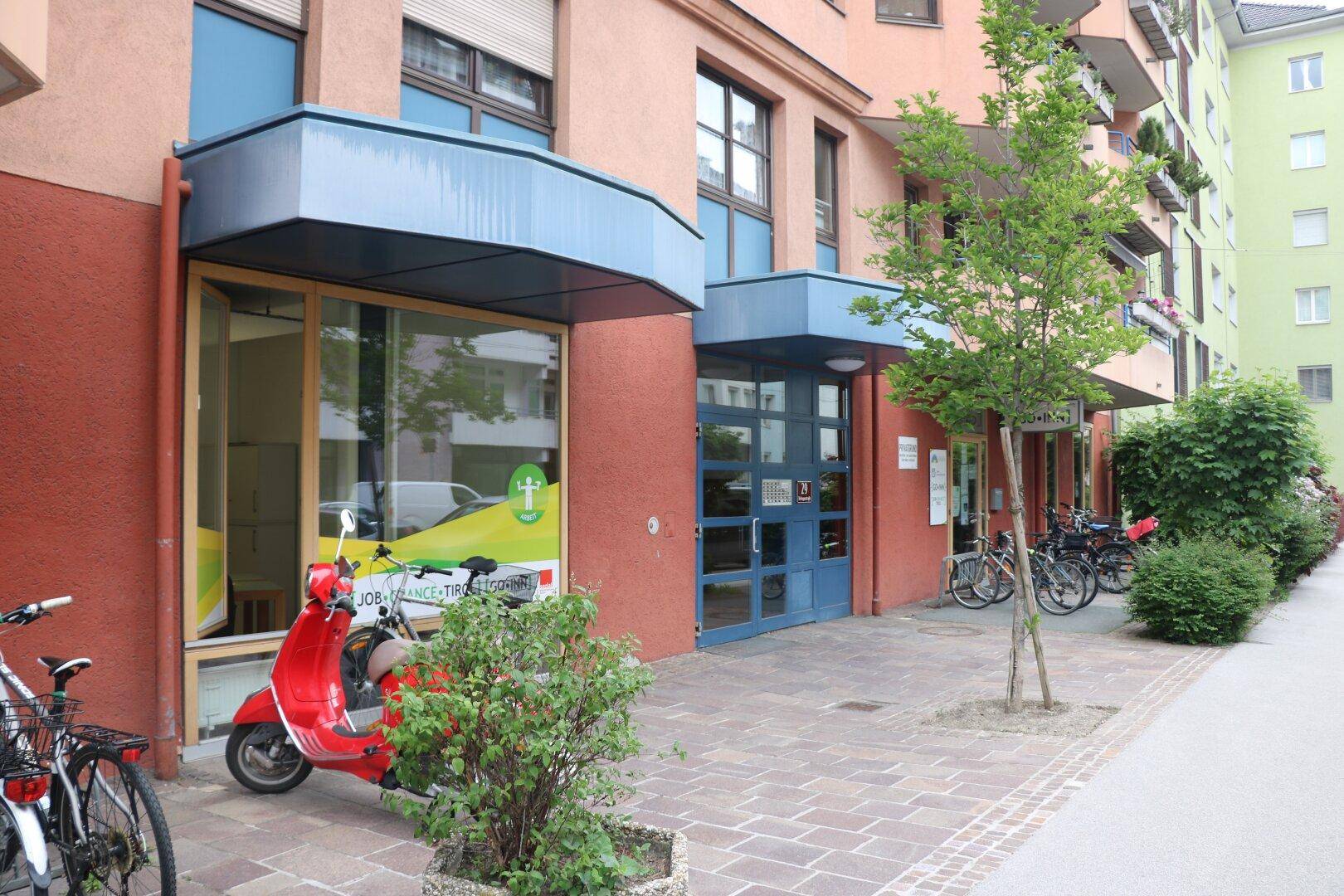 Eingangsbereich eines Gebäudes mit Schaufenstern, einer roten Vespa und Fahrradständern.