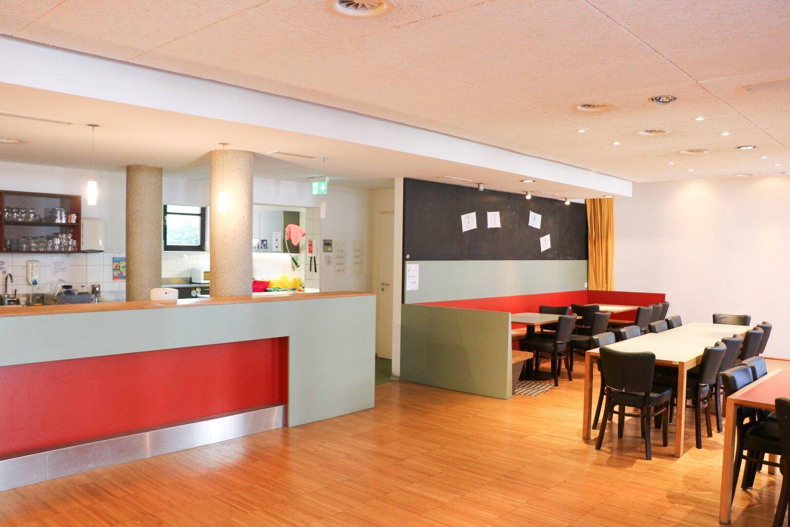 Großer Speisesaal mit Holzboden, langen Tischen und einer Theke im Hintergrund.