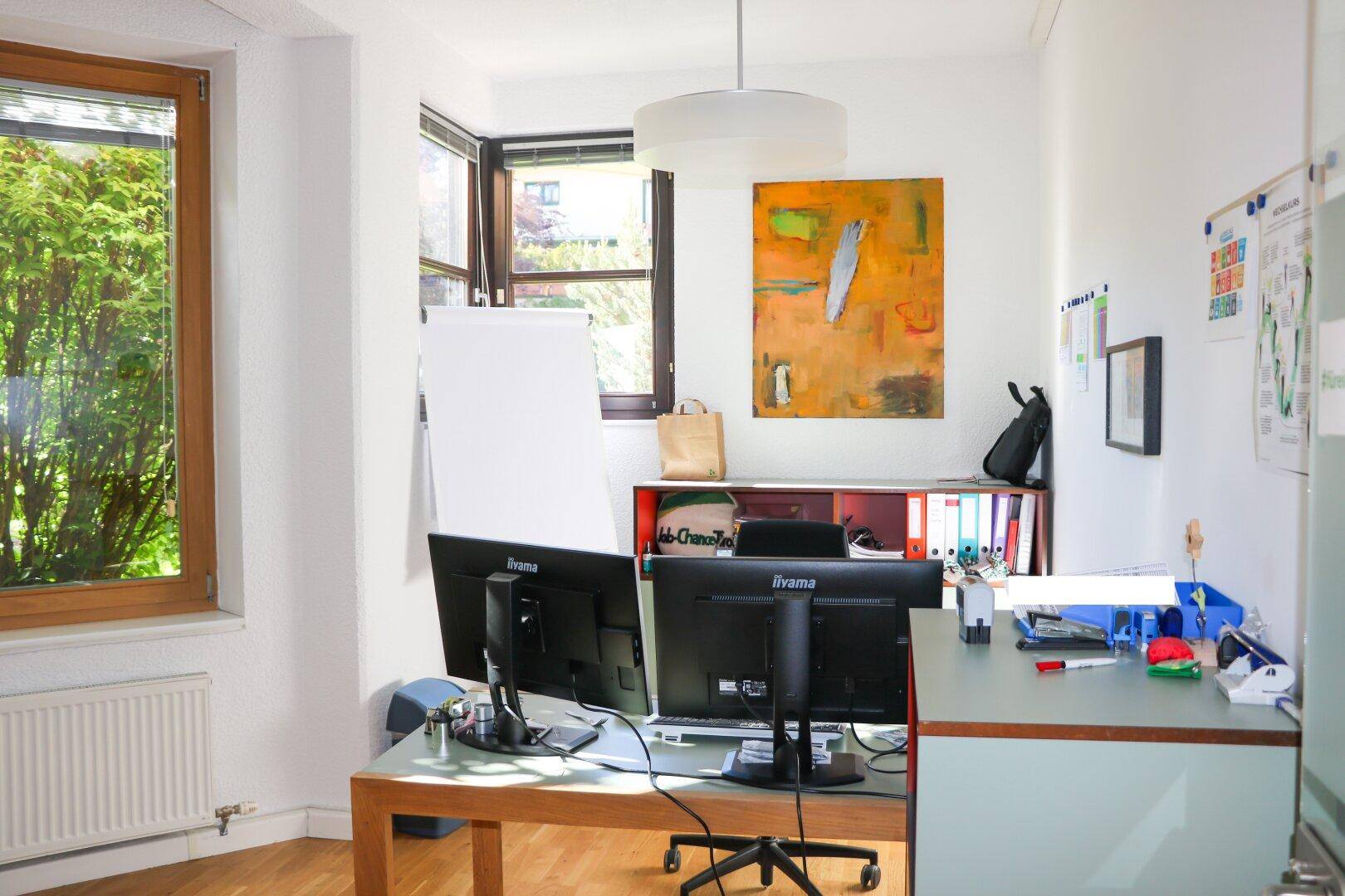 Helles Büro mit zwei Computermonitoren, einem Schreibtisch und großen Fenstern mit Grünblick.