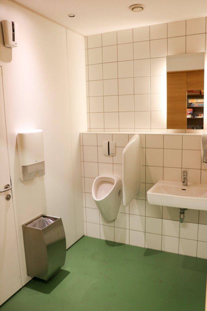 Saubere Toilette mit weißen Fliesen, Urinal, Waschbecken und grünem Boden.