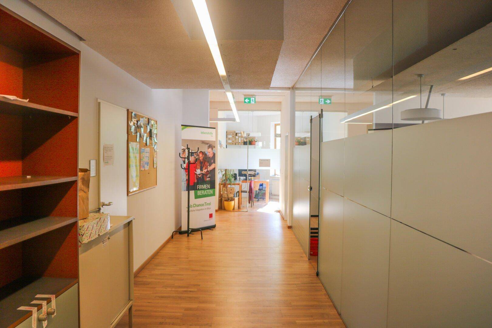 Heller Bürogang mit Glaswänden und Holzfußboden, der zu weiteren Arbeitsbereichen führt.