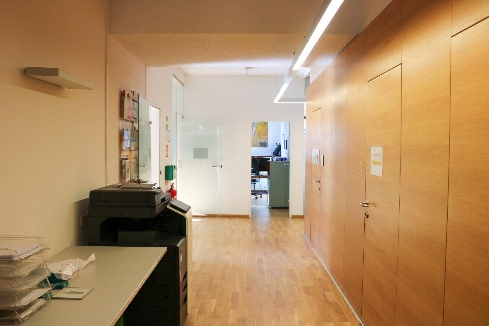 Langer Flur mit Holzverkleidung, einem Drucker und Blick in einen weiteren Bürobereich.
