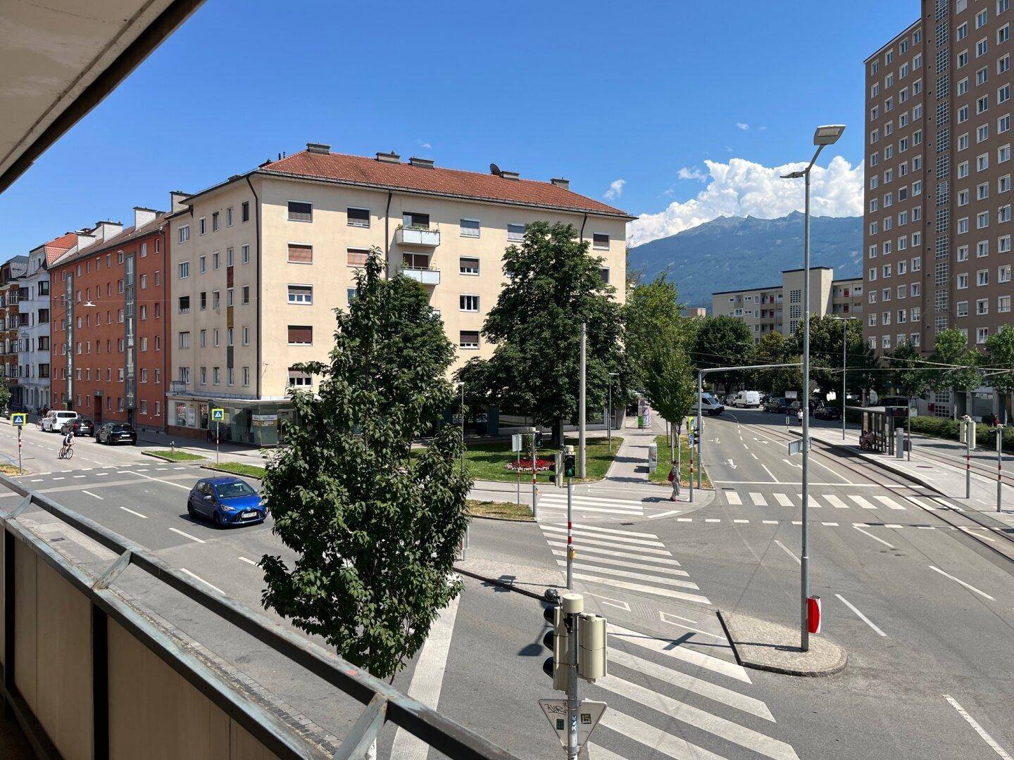 Weitblick über eine belebte Stadtkreuzung mit Gebäuden, Bäumen und Bergen im Hintergrund.