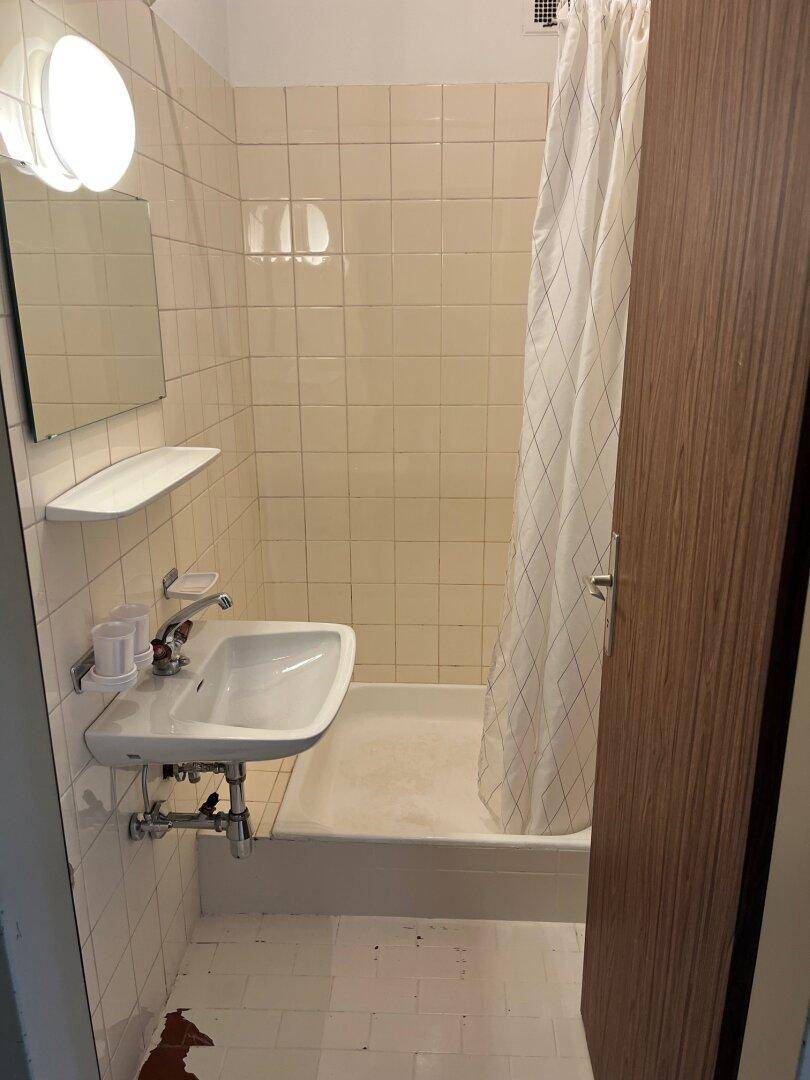 Badezimmer mit beigefarbenen Fliesen, Waschbecken, Spiegel und Duschbereich mit Vorhang.