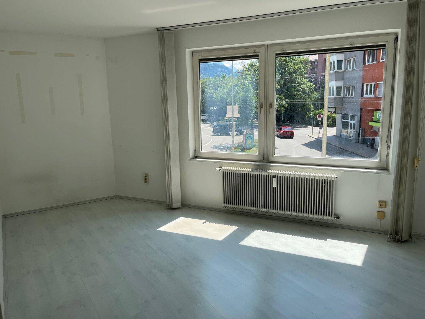 Helles Zimmer mit großem Fenster, Laminatboden und Blick auf die Straße und Berge.