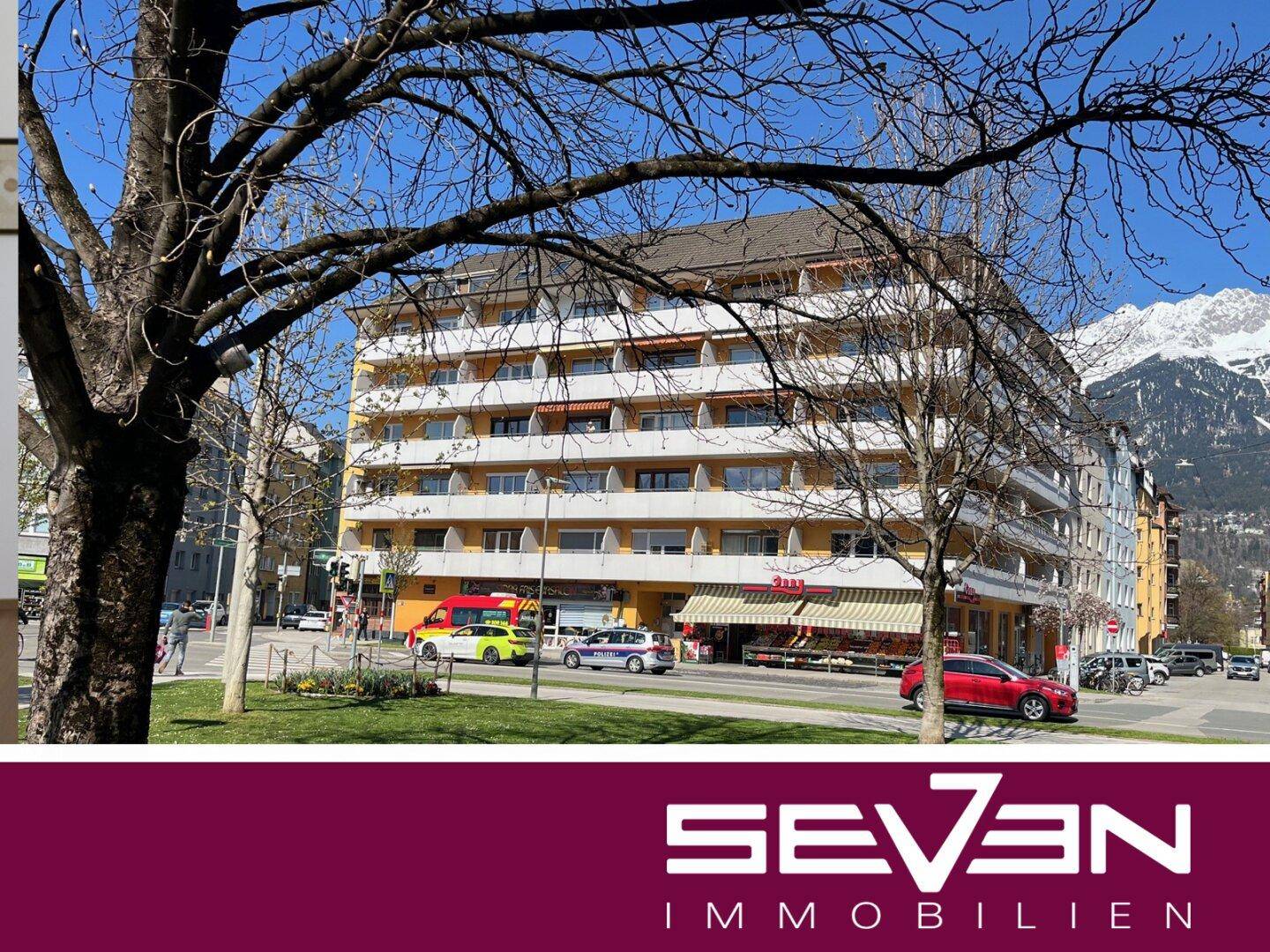 Mehrstöckiges Wohngebäude mit gelber Fassade, Balkonen und Geschäften im Erdgeschoss, umgeben von Bäumen und Bergen im Hintergrund.