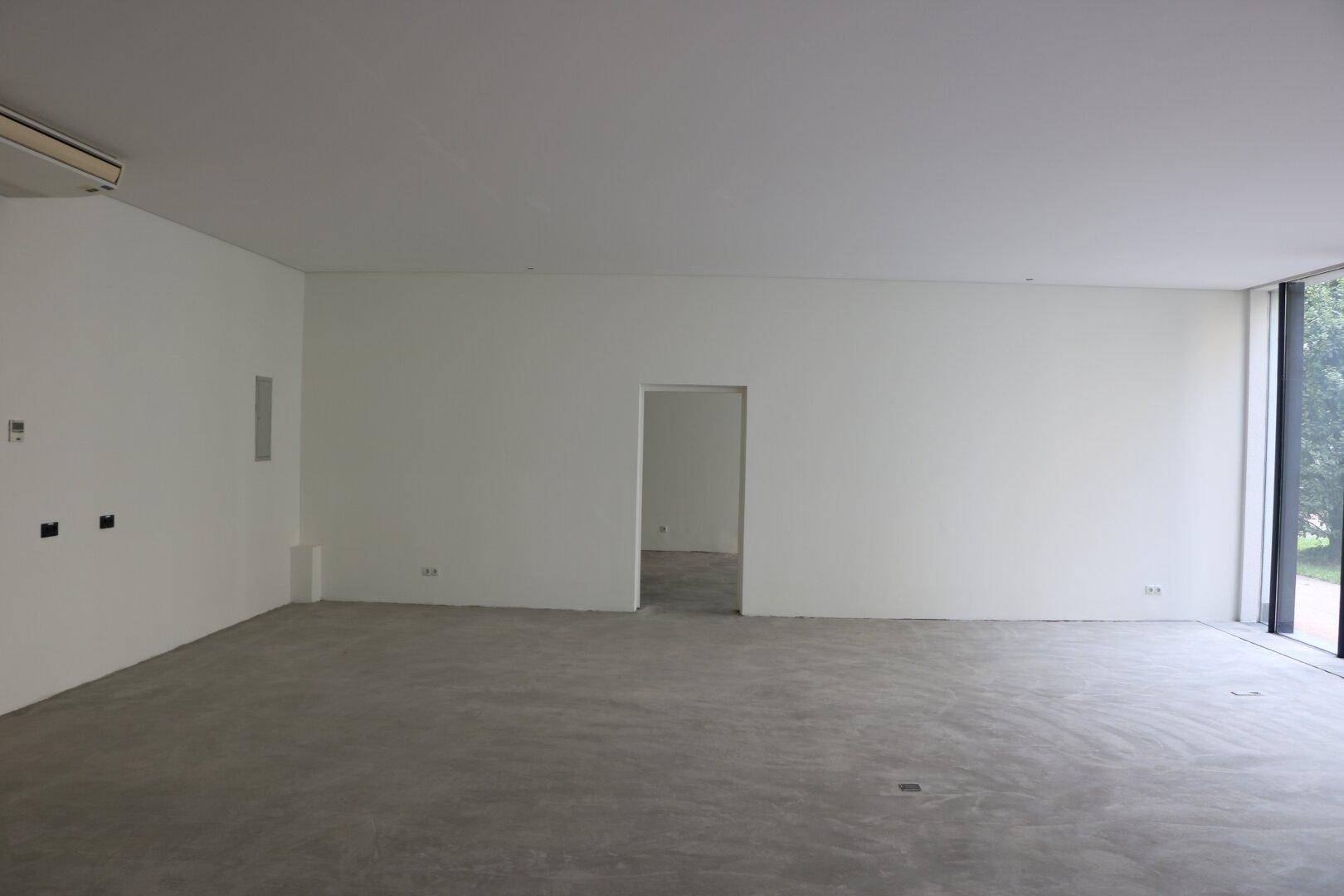 Großer, leerer Raum mit weißen Wänden, Betonboden und Klimaanlage an der Decke.