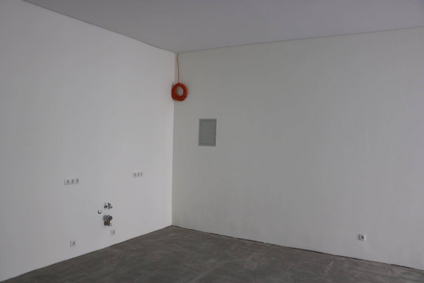 Leerer Raum mit weißen Wänden, Betonboden und sichtbaren Anschlüssen für Installationen.