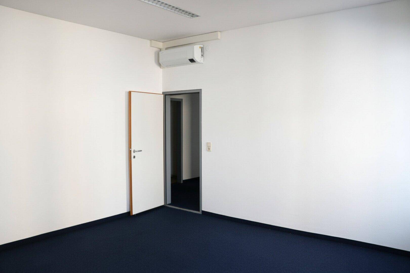 Leeres Büro mit blauem Teppichboden, weißen Wänden, einer Klimaanlage und einer offenen Tür.