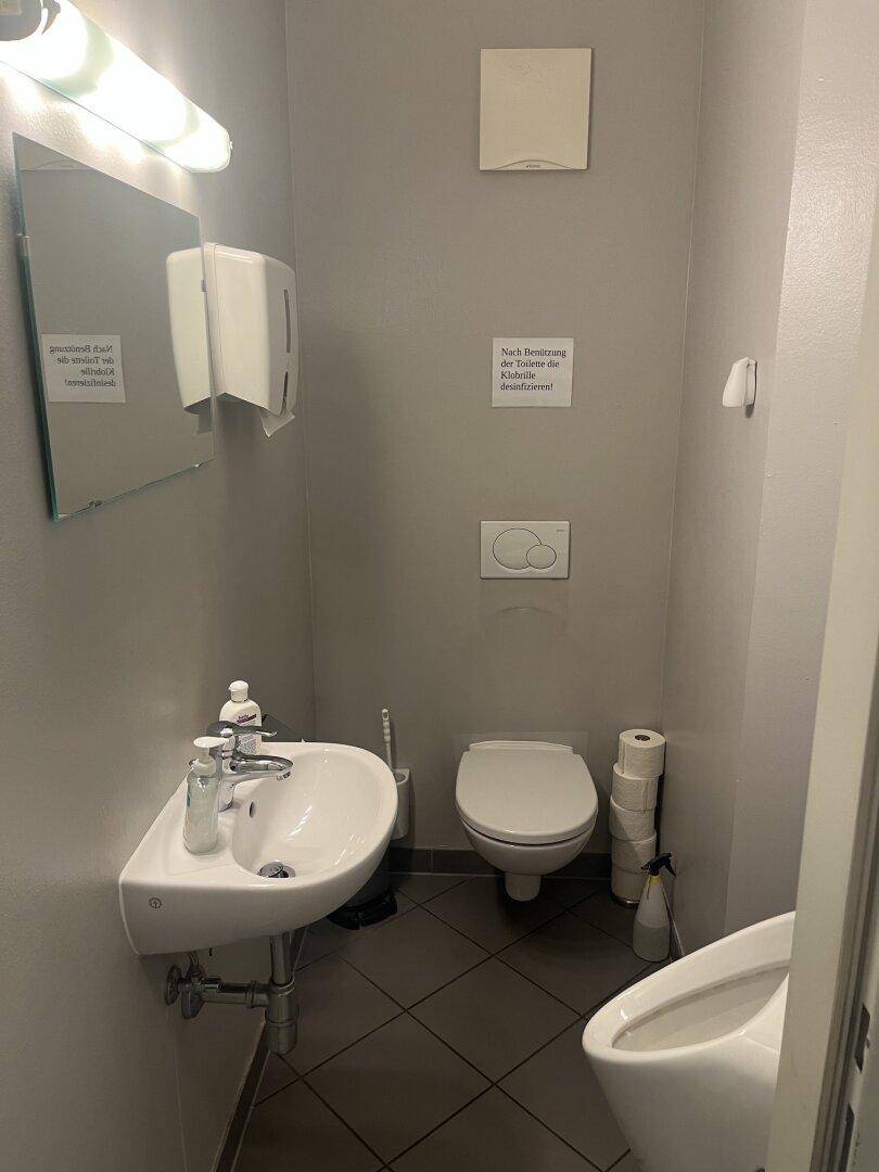Kleine Gästetoilette mit Waschbecken, WC, Urinal und dunklen Fliesen.