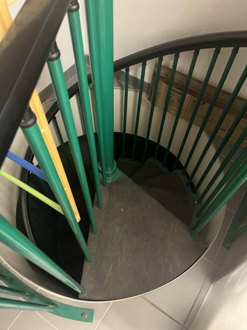 Eine enge Wendeltreppe mit grünen Geländerstäben und dunklen Stufen, die nach unten führt.