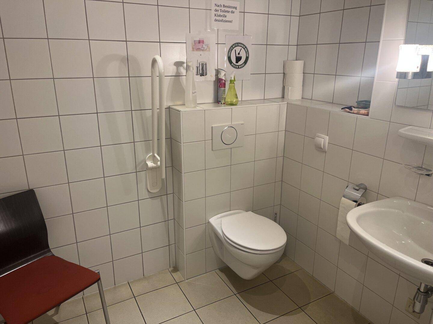 Barrierefreie Toilette mit Haltegriff, WC, Waschbecken und weißen Wandfliesen.