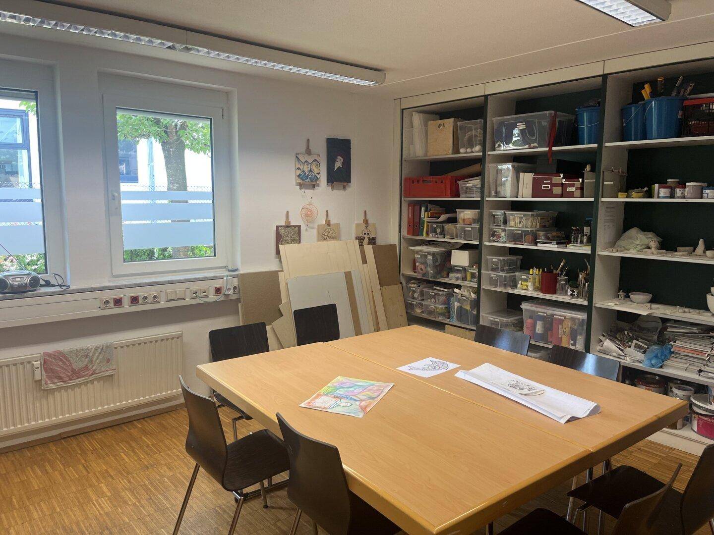 Kreativraum oder Werkstatt mit großem Tisch, Regalen voller Materialien und Fenstern.