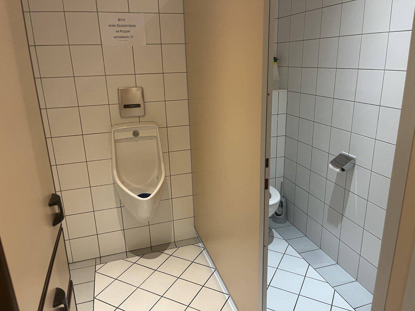 Herrentoilette mit Urinal, WC und weißen Fliesen an Wänden und Boden.