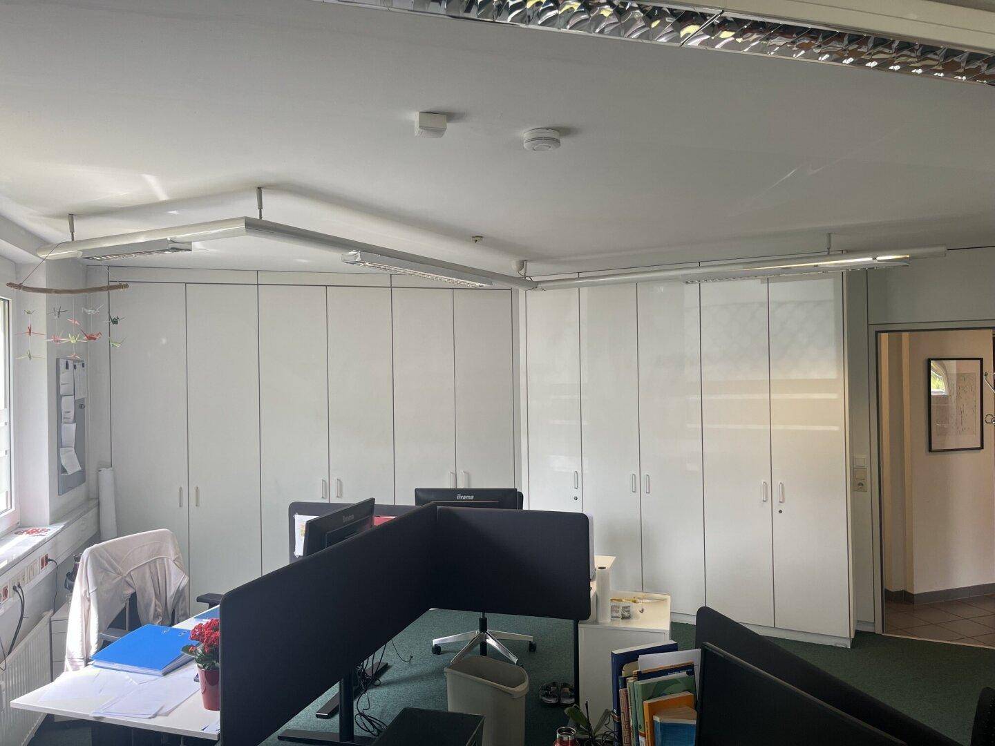 Großraumbüro mit weißen Schränken entlang der Wand und mehreren Arbeitsplätzen.