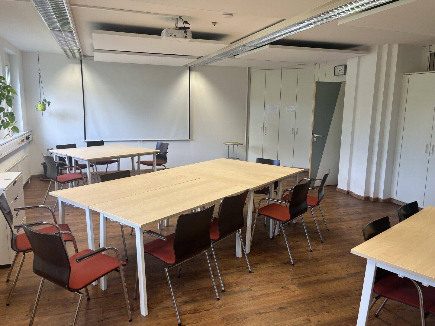 Konferenzraum mit Projektionsfläche, mehreren Tischen und Stühlen sowie Einbauschränken.