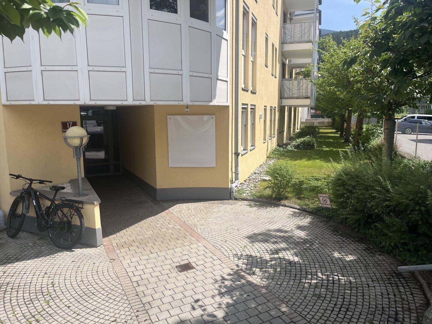 Eingangsbereich eines Mehrfamilienhauses mit gepflastertem Weg und Fahrradabstellplatz.