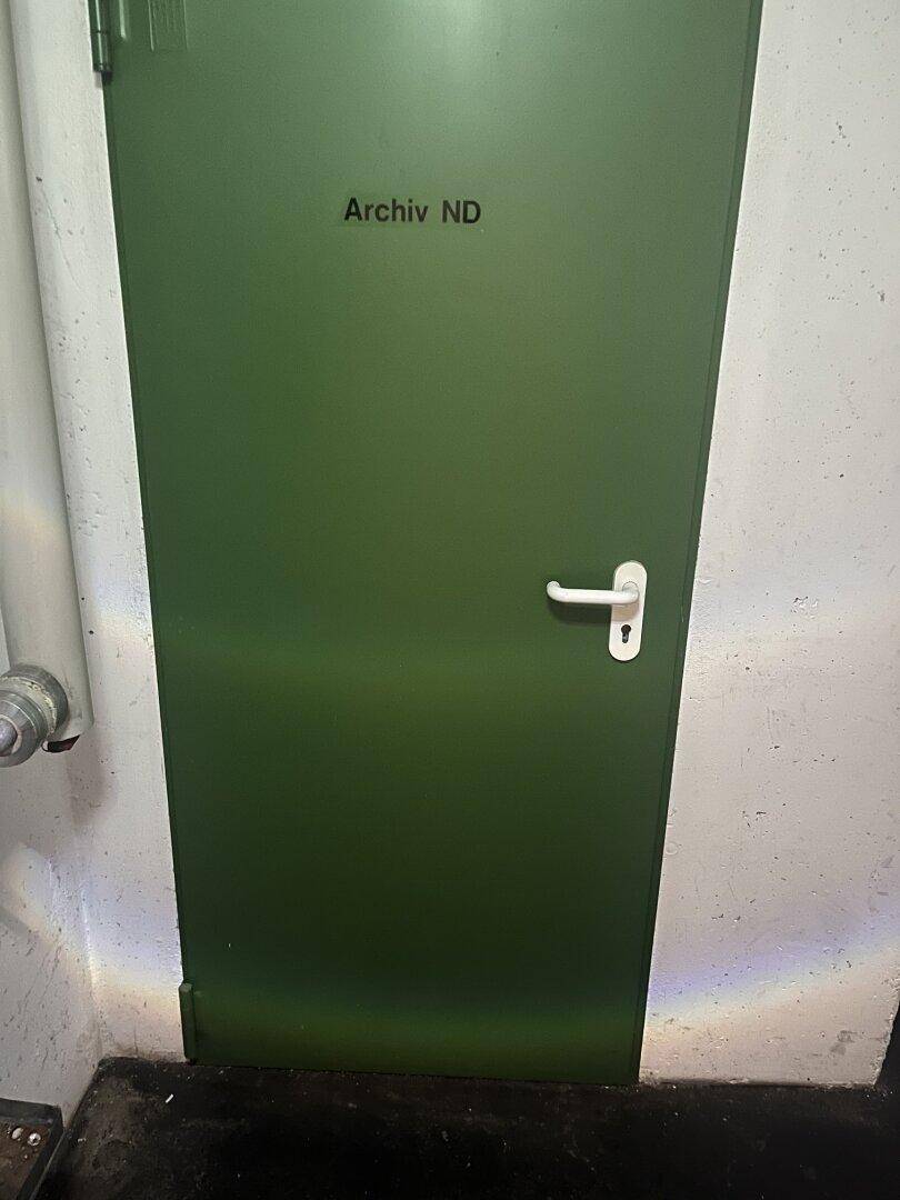 Eine grüne Tür mit der Aufschrift "Archiv ND" in einem Kellerbereich mit rauen Wänden.