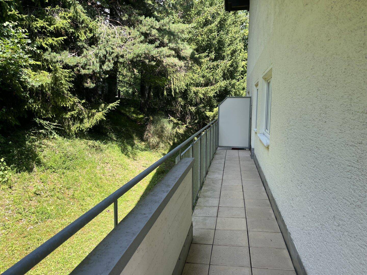 Schmaler Balkon mit Fliesenboden und einfacher Brüstung, umgeben von viel Grün.