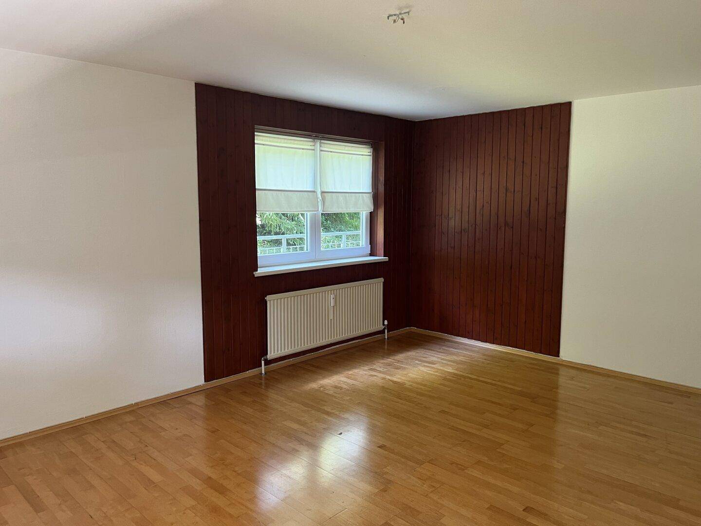 Leeres Zimmer mit Holzboden, einer Holzwand und Fenster mit Blick ins Grüne.