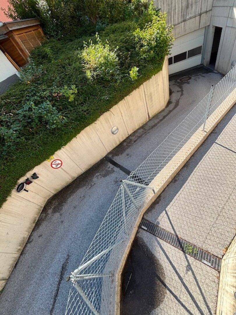 Draufsicht auf die Zufahrt zur Tiefgarage mit Betonmauern, Hecke und Metallzaun.