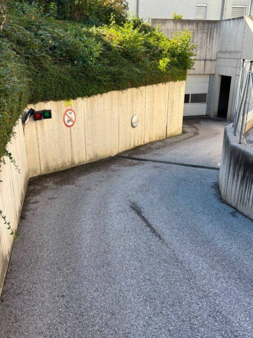 Einfahrt zur Tiefgarage mit Betonwänden, Hecke und Verkehrsschildern.