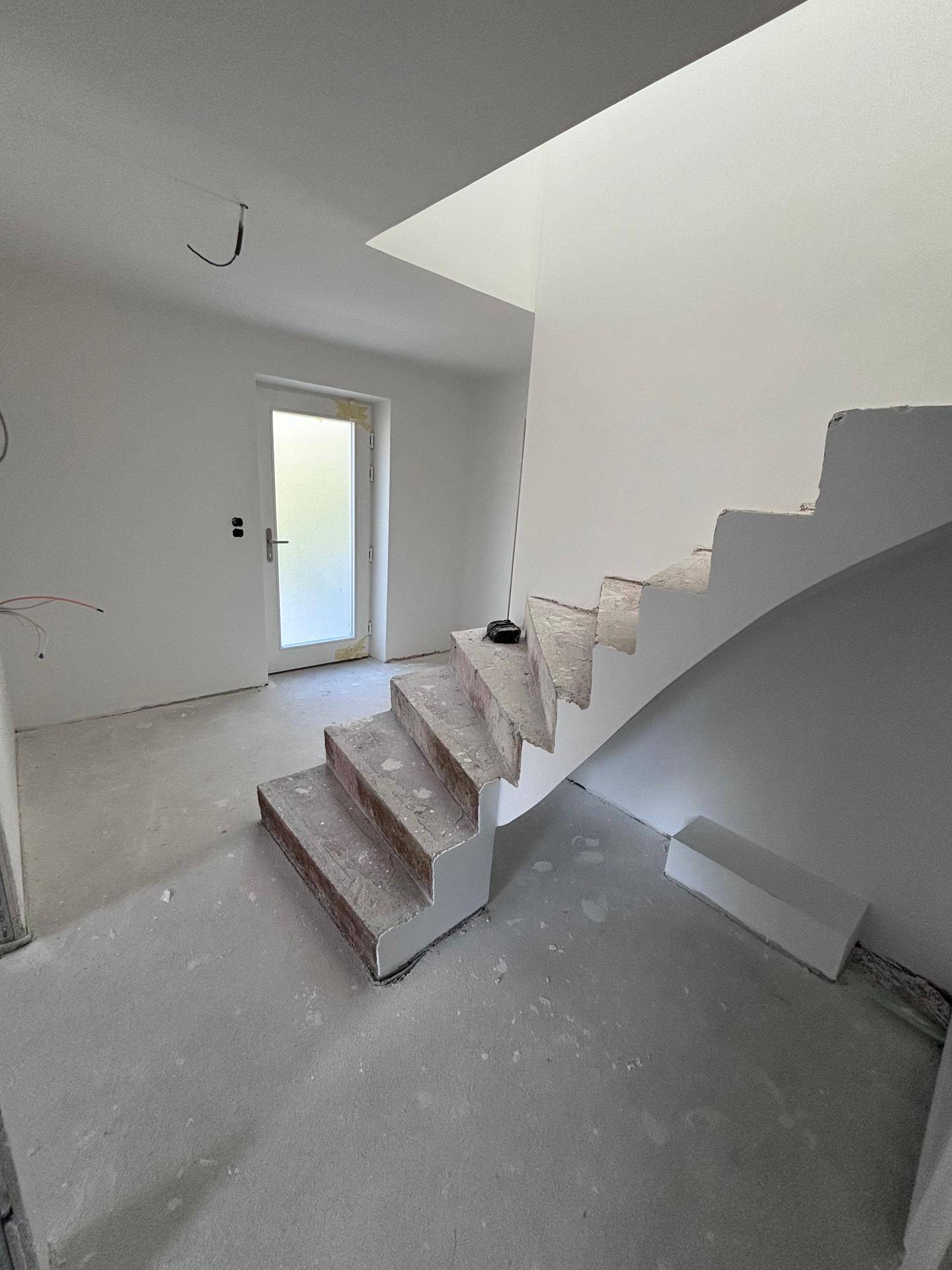 Eingangsbereich im Rohbau mit offener Treppe und weißer Tür, bereit für den Innenausbau.