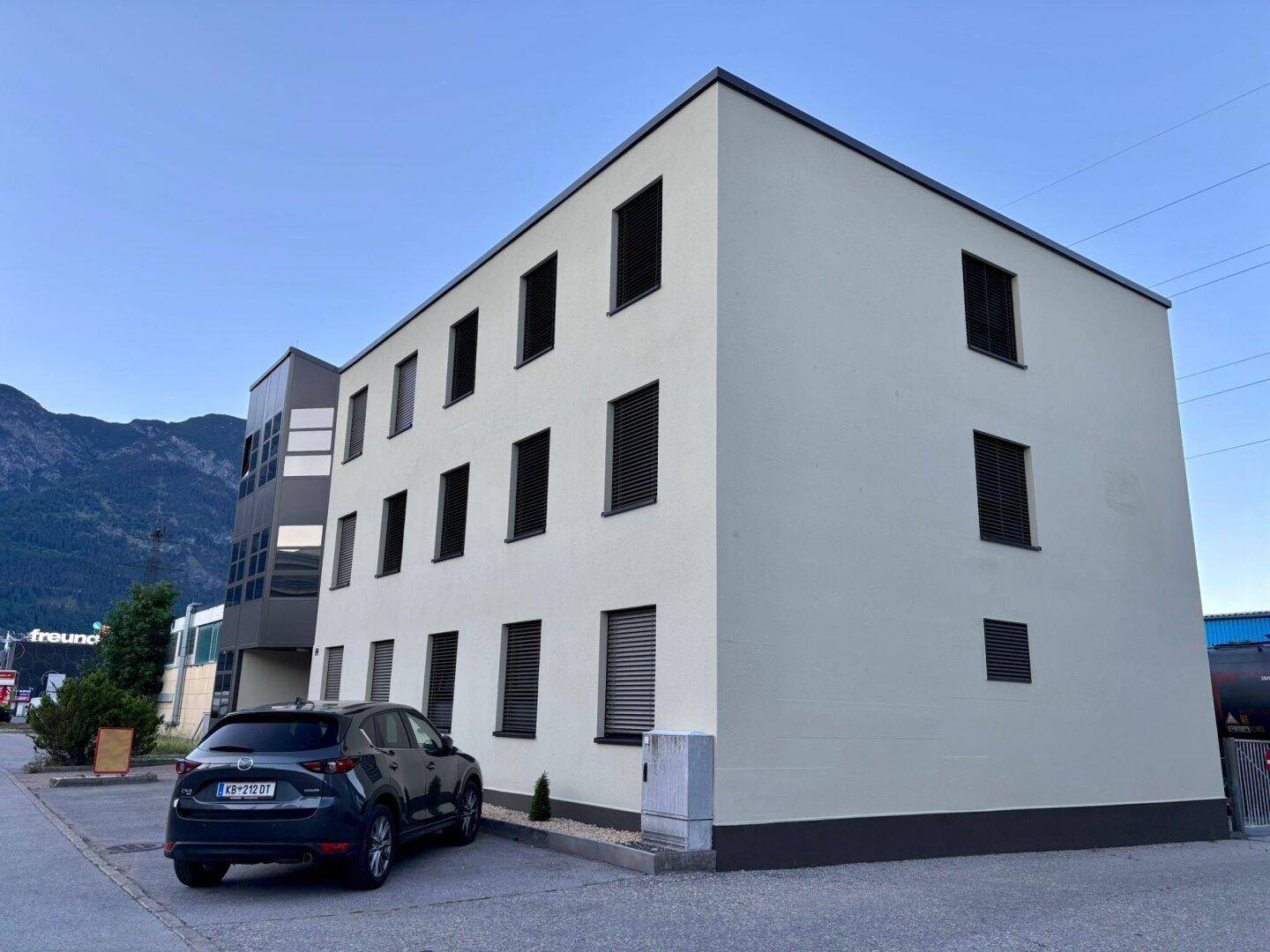 Gesamtansicht des modernen Gebäudes mit geparktem Auto und Bergen im Hintergrund unter klarem Himmel.