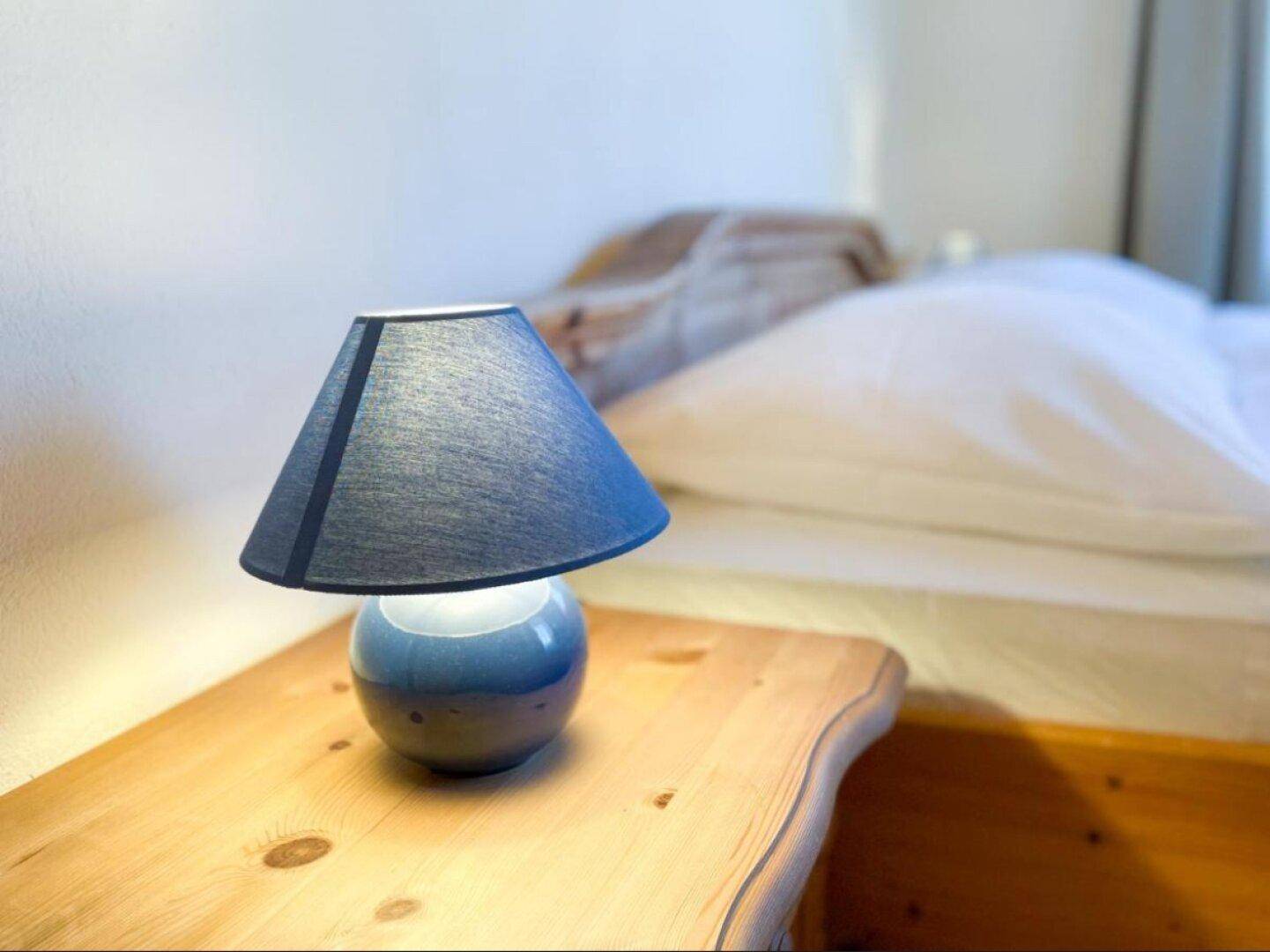 Gemütliche Nachttischlampe auf einem Holztisch neben einem Bett in einem hellen Schlafzimmer.