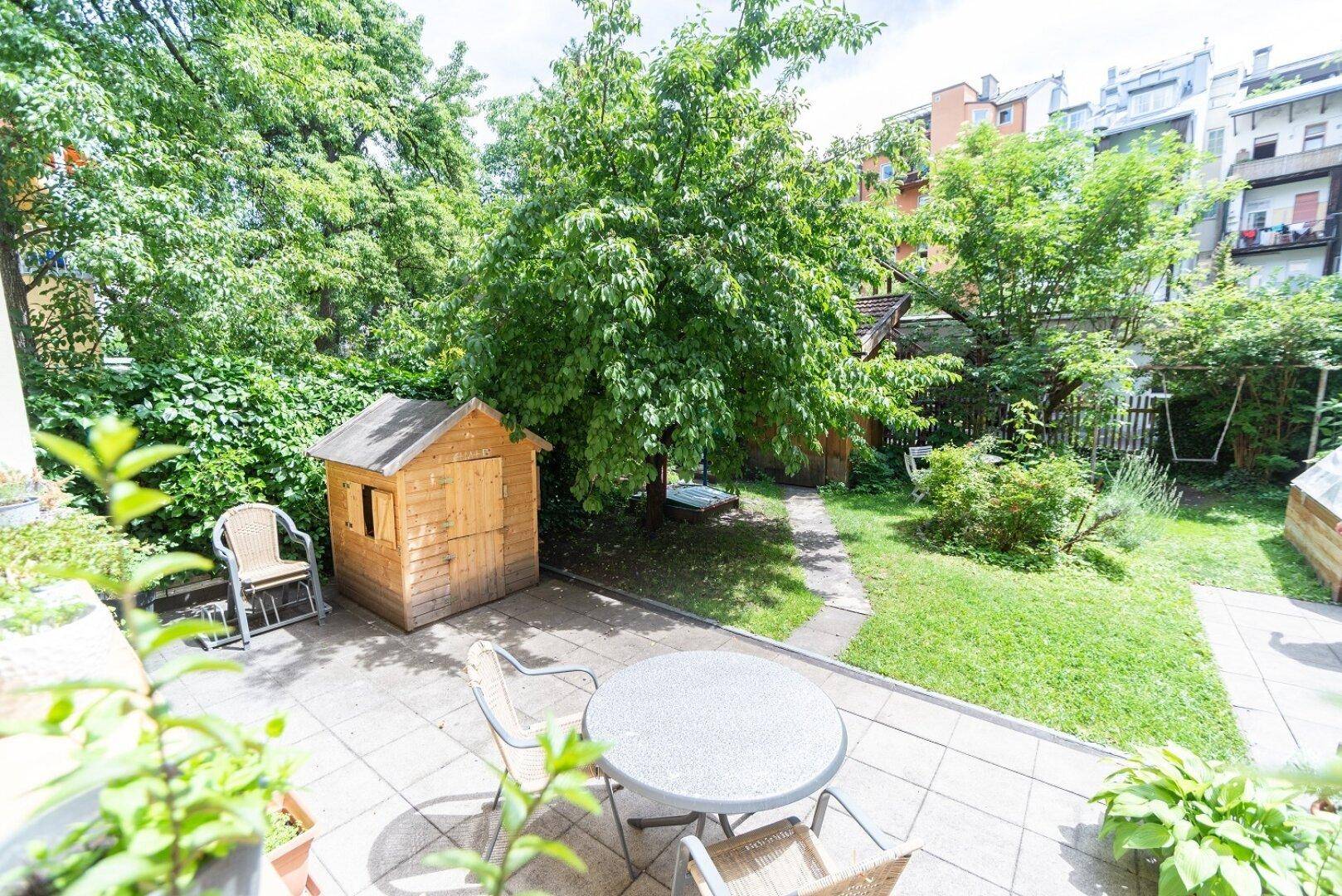 Grüner Garten mit gepflasterter Terrasse, Kinderspielhaus und üppiger Vegetation.