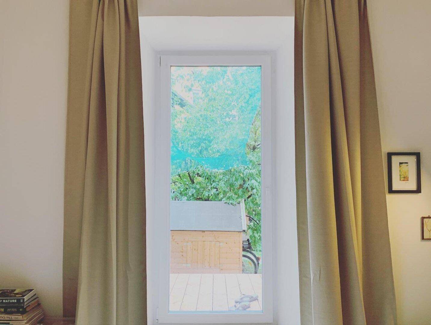 Fensterblick auf einen grünen Garten mit Gartenhaus und Holzboden im Freien.
