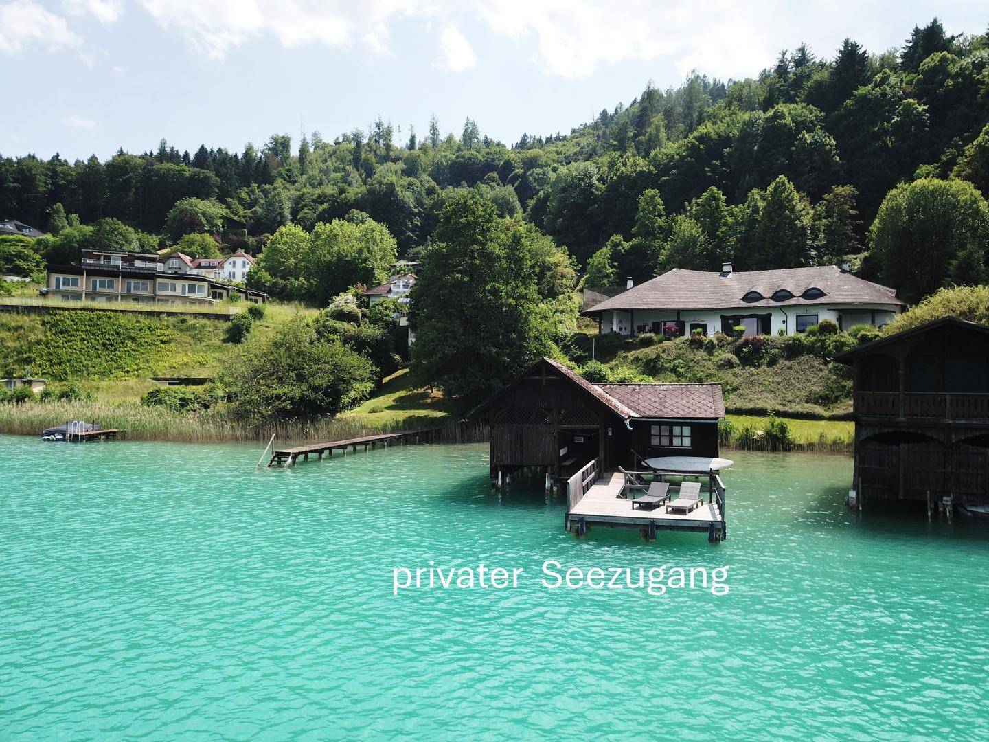 Privater Seezugang mit einem Holzsteg und einem Bootshaus direkt am türkisfarbenen Wasser des Sees.