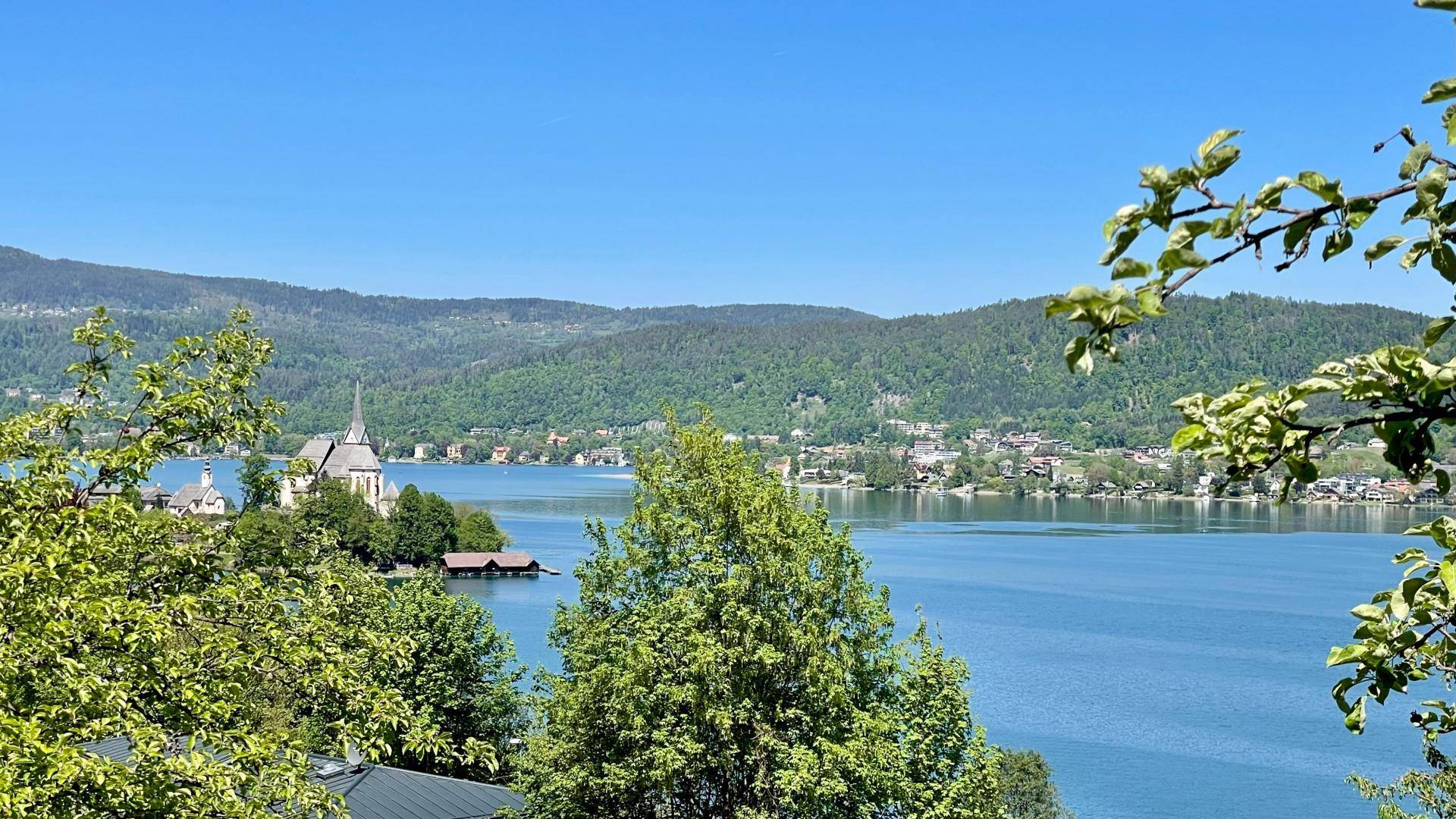 Panoramablick über den See mit der Inselkirche und den grünen, bewaldeten Bergen im Hintergrund.