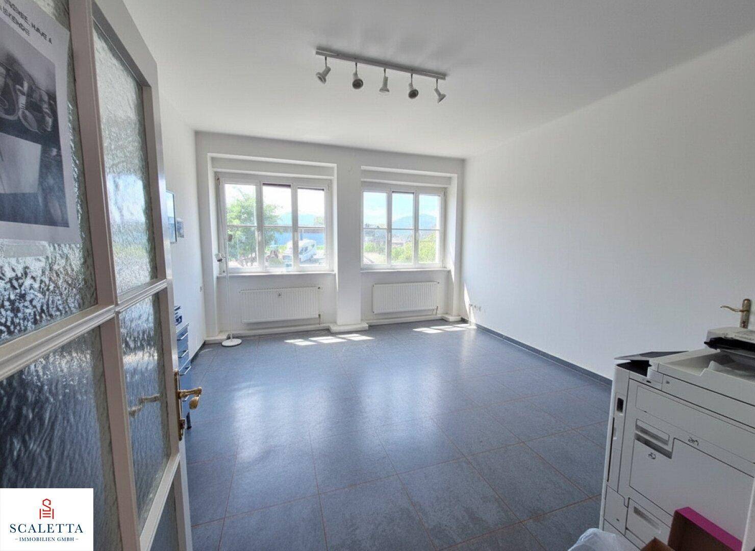Heller Büroraum mit zwei großen Fenstern, die viel Tageslicht hereinlassen, und grauen Fliesen.