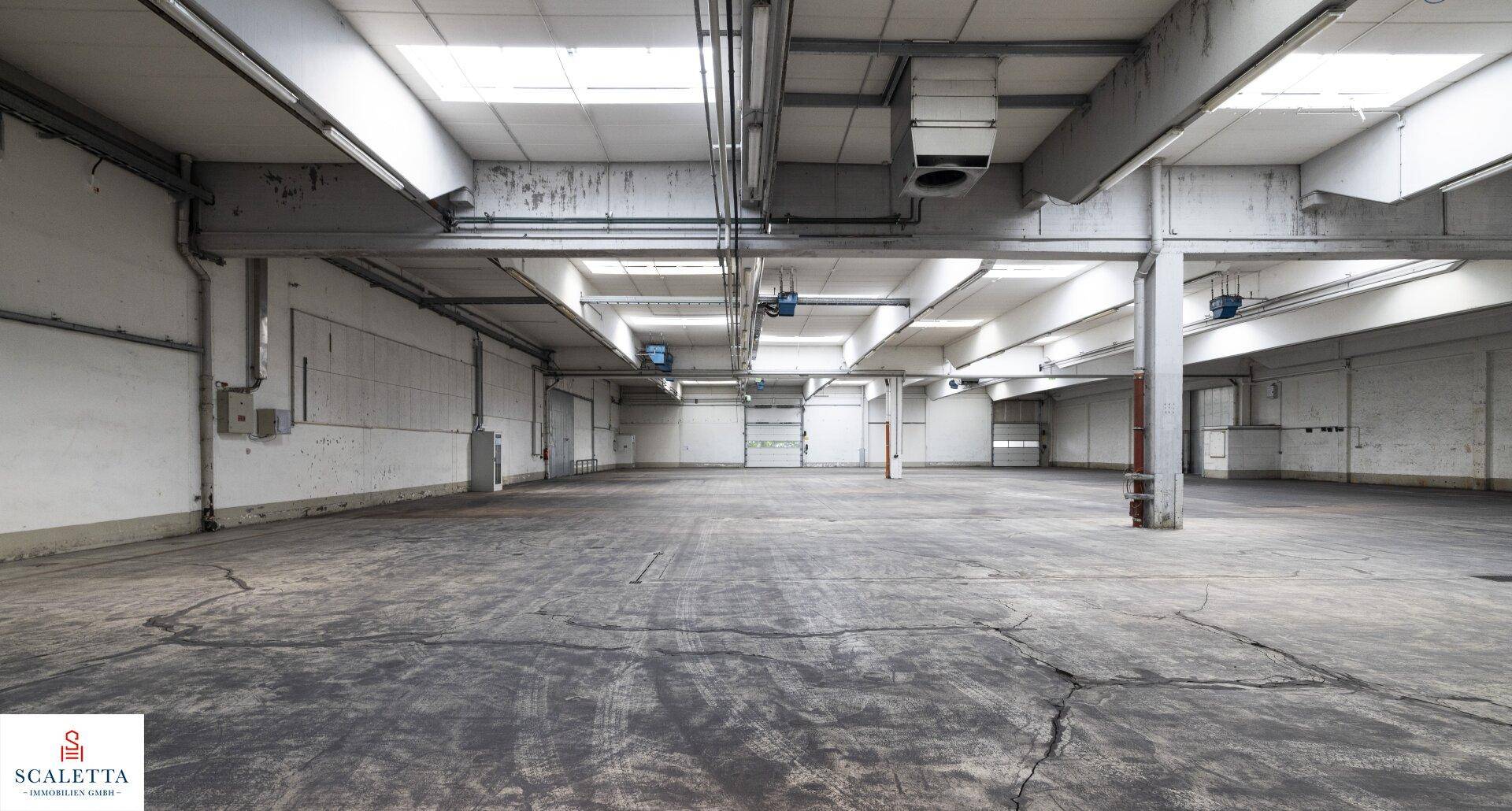 Industrielle Lagerhalle mit sichtbaren Lüftungsanlagen, Betonboden und Oberlichtern für natürliche Beleuchtung.