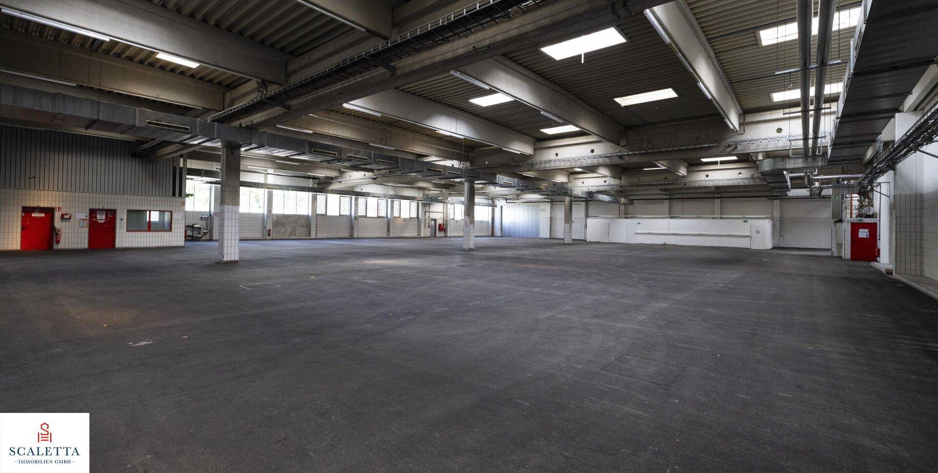 Große Lagerhalle mit roten Türen und Fenstern entlang einer Wand, die Tageslicht hereinlassen.