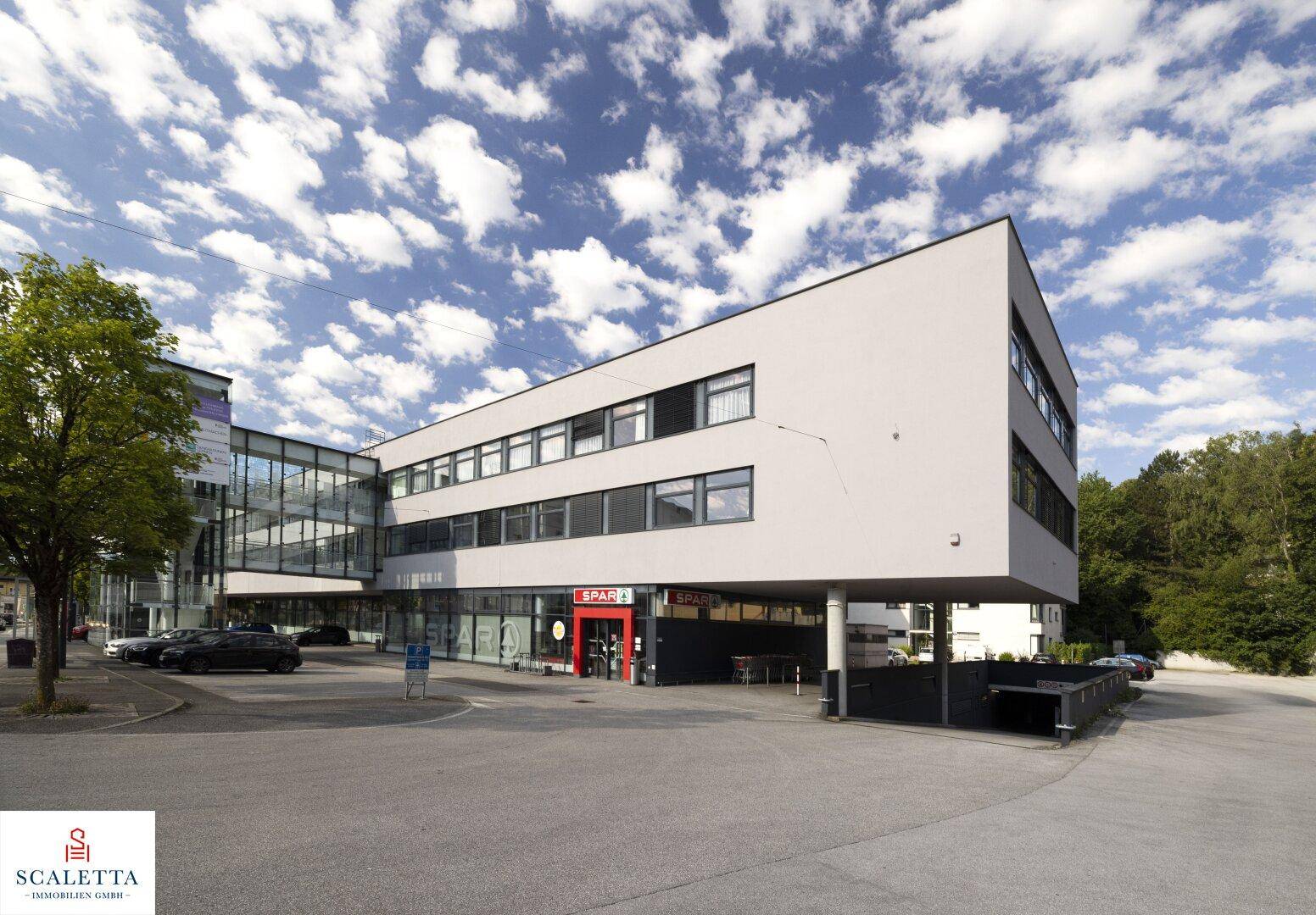 Modernes Geschäftsgebäude mit Supermarkt im Erdgeschoss und Parkplätzen unter blauem Himmel.